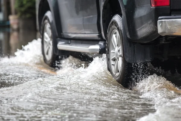 Rear tire and wheel of a dark SUV splashing through deep water on a flooded road.