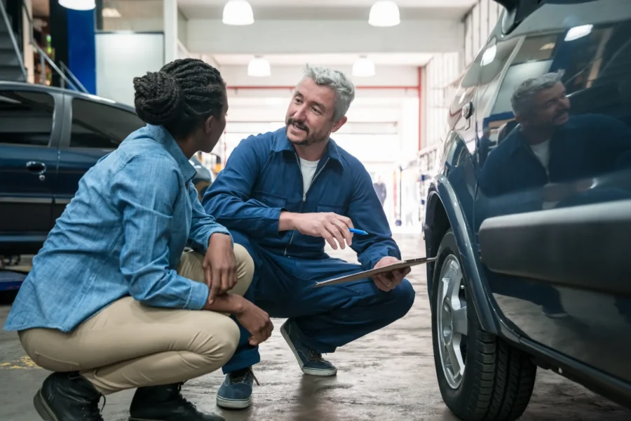 A mechanic in blue coveralls holding a clipboard crouches beside an SUV's front tire, talking with a woman customer crouched next to him in a repair shop bay.