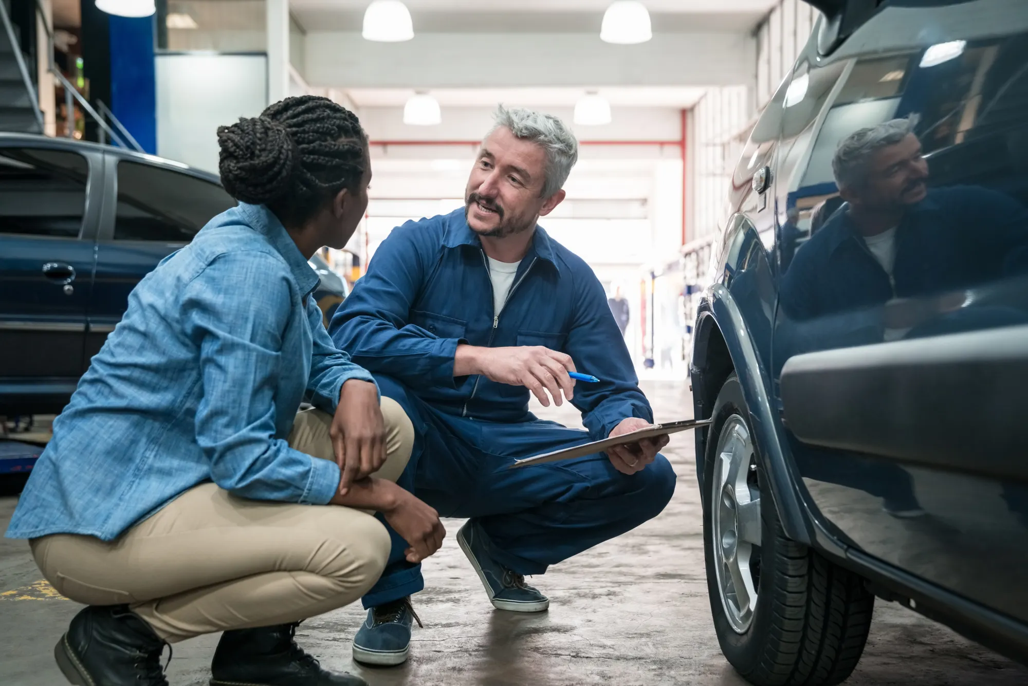 A mechanic in blue coveralls holding a clipboard crouches beside an SUV's front tire, talking with a woman customer crouched next to him in a repair shop bay.